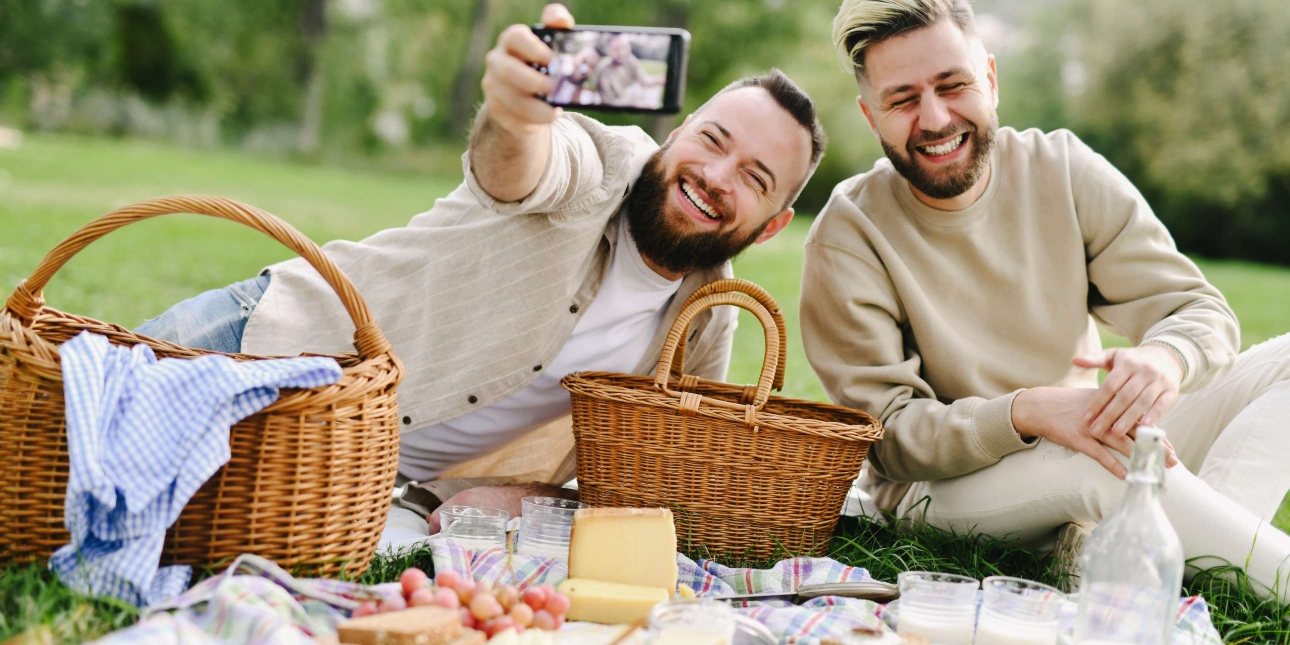 Two white men with beards sit laughing outdoors on a picnic blanket. The man on the left is taking a selfie of them both surrounded by picnic baskets, fruit, bread and drinks.