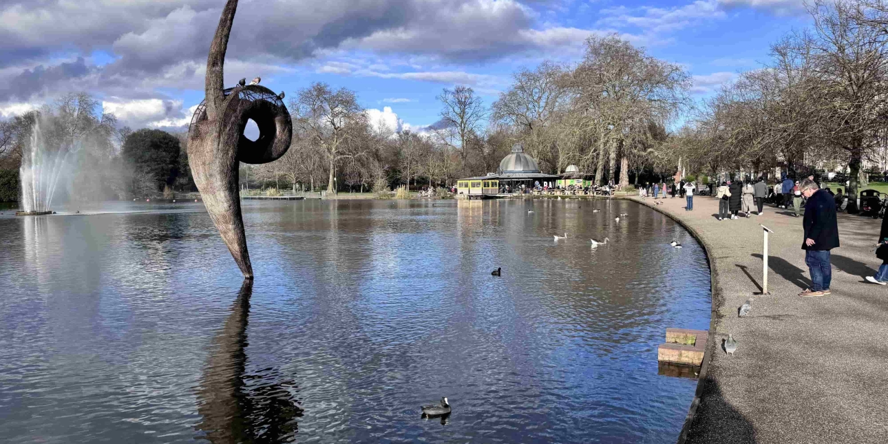 The boating at Victoria Park. White and grey clouds in the sky are reflected into the lake, which is edged with a tree-lined concrete path. People are walking past. A sculpture emerges from the lake. Its form is like a curvy line with a small loop in