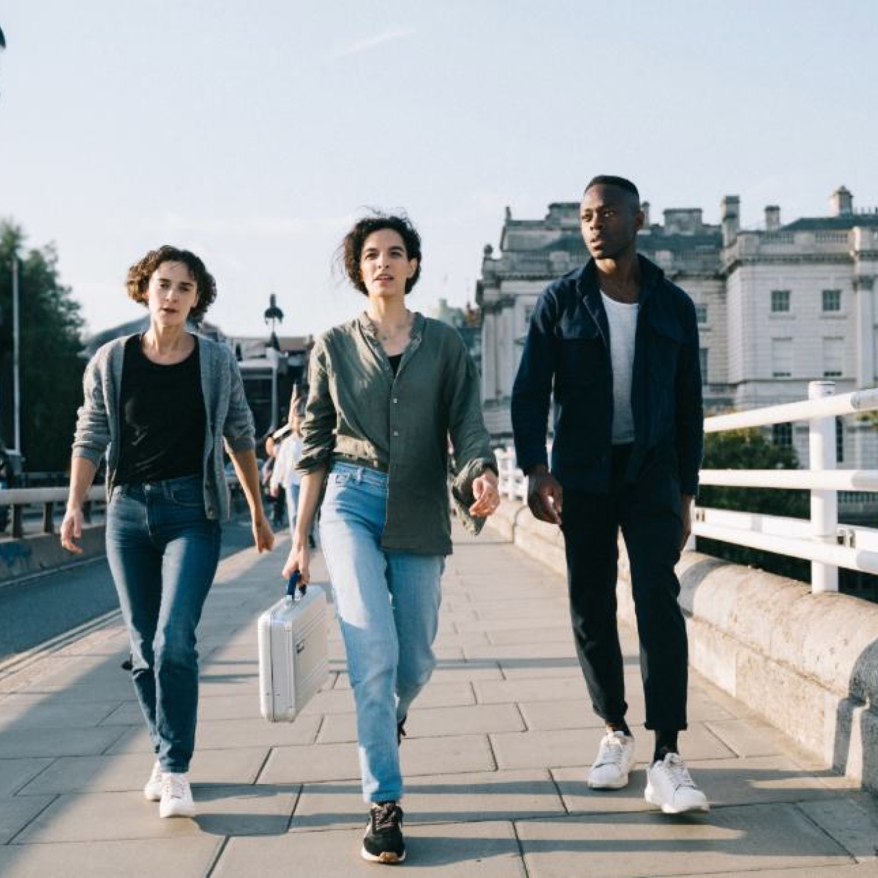 Three people - two women and one man - striding across a bridge in London. The woman in the middle is holding a metallic briefcase.