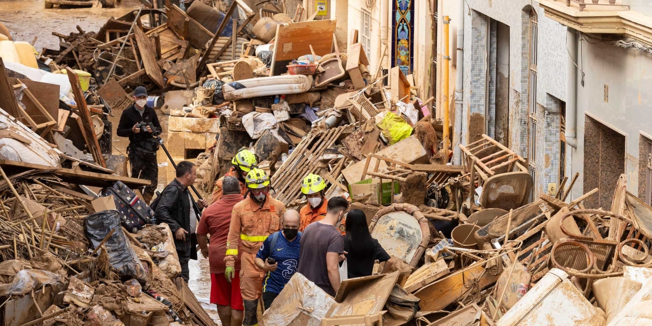 A group of including rescuers walking through a piles of damaged, mud covered furniture and debris on a street