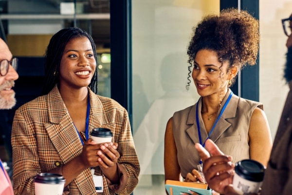 A group of four smiling colleagues of different ages stood holding coffee cups: a white man with grey beard, a Black woman with check jacket, a Black woman wearing an sleeveless top, and a Black man wearing a brown shirt.