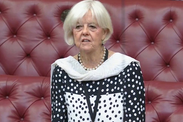 Baroness Hayter - a white woman with grey hair who is wearing a monochrome spotted top - is stood in the House of Lords chamber in front of red benches.