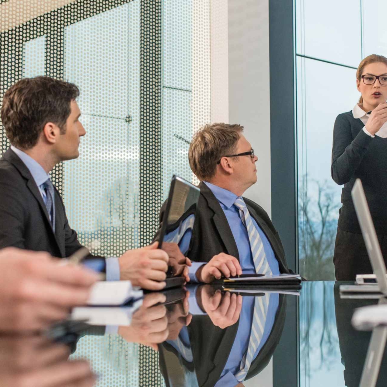 In a modern boardroom a solitary white business woman gives a presentation in front of three white men in suits.
