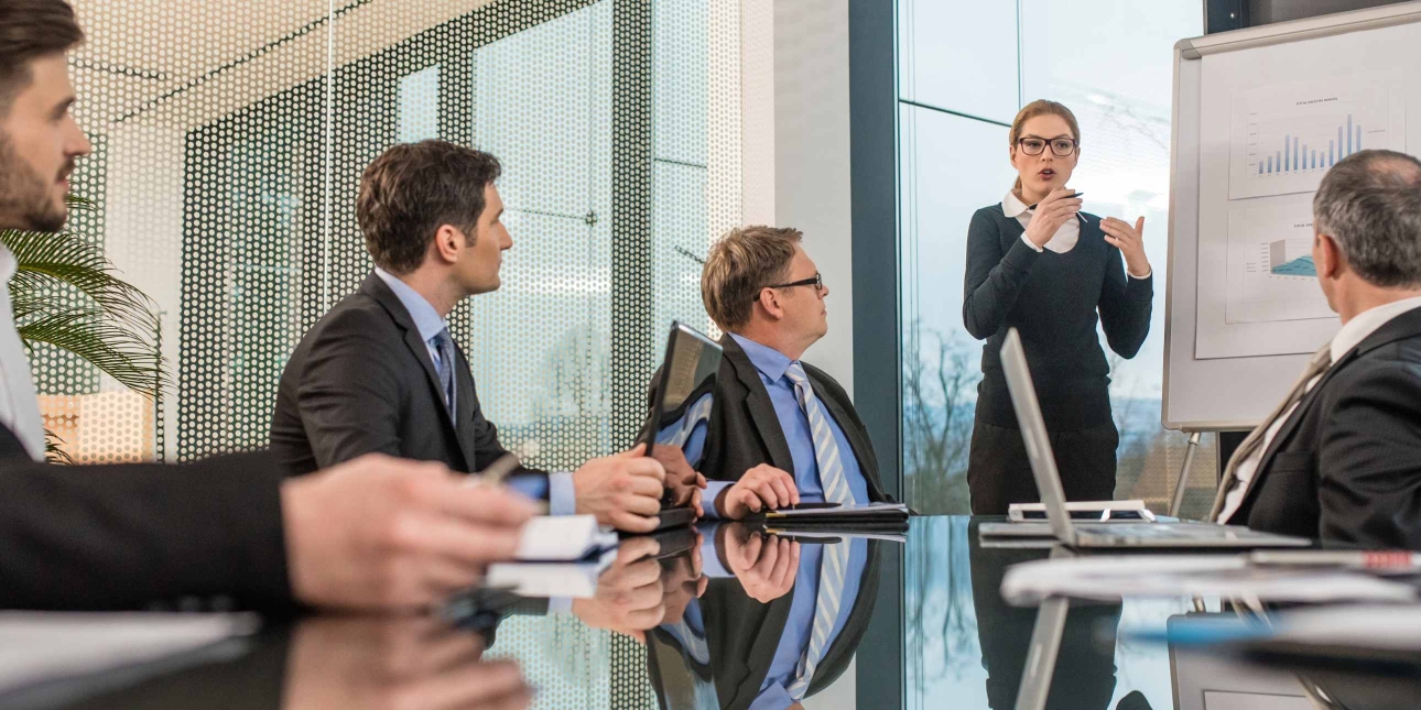 In a modern boardroom a solitary white business woman gives a presentation in front of three white men in suits.