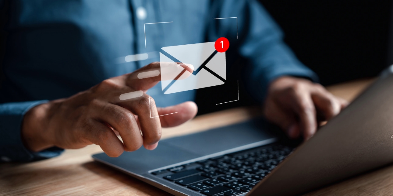 A colour photograph of an unidentified person wearing a blue shirt pointing at his laptop screen. Super-imposed over the photo is a computer generated white icon of an envelope.