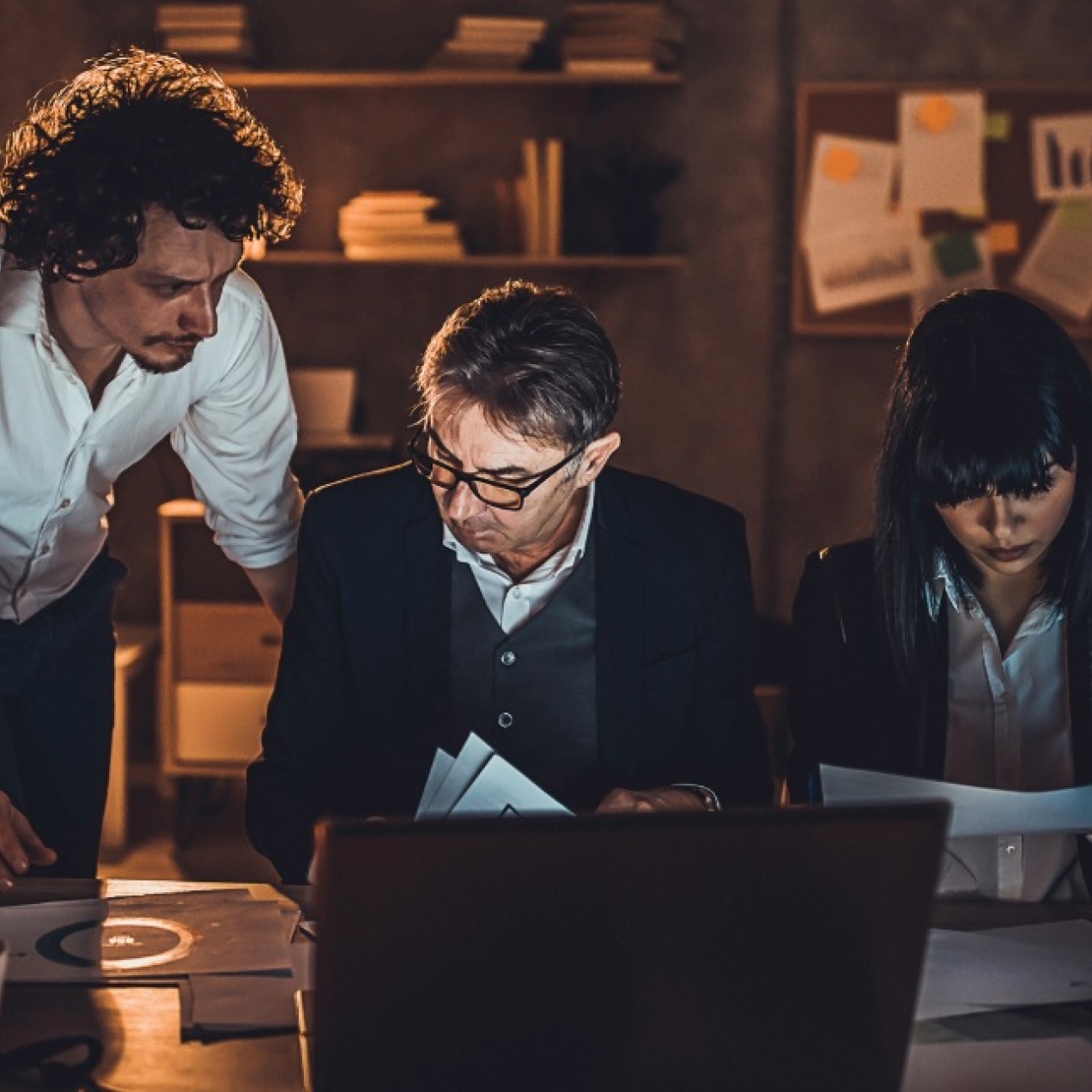 In a dark office, three colleagues work late into the night around a desk.