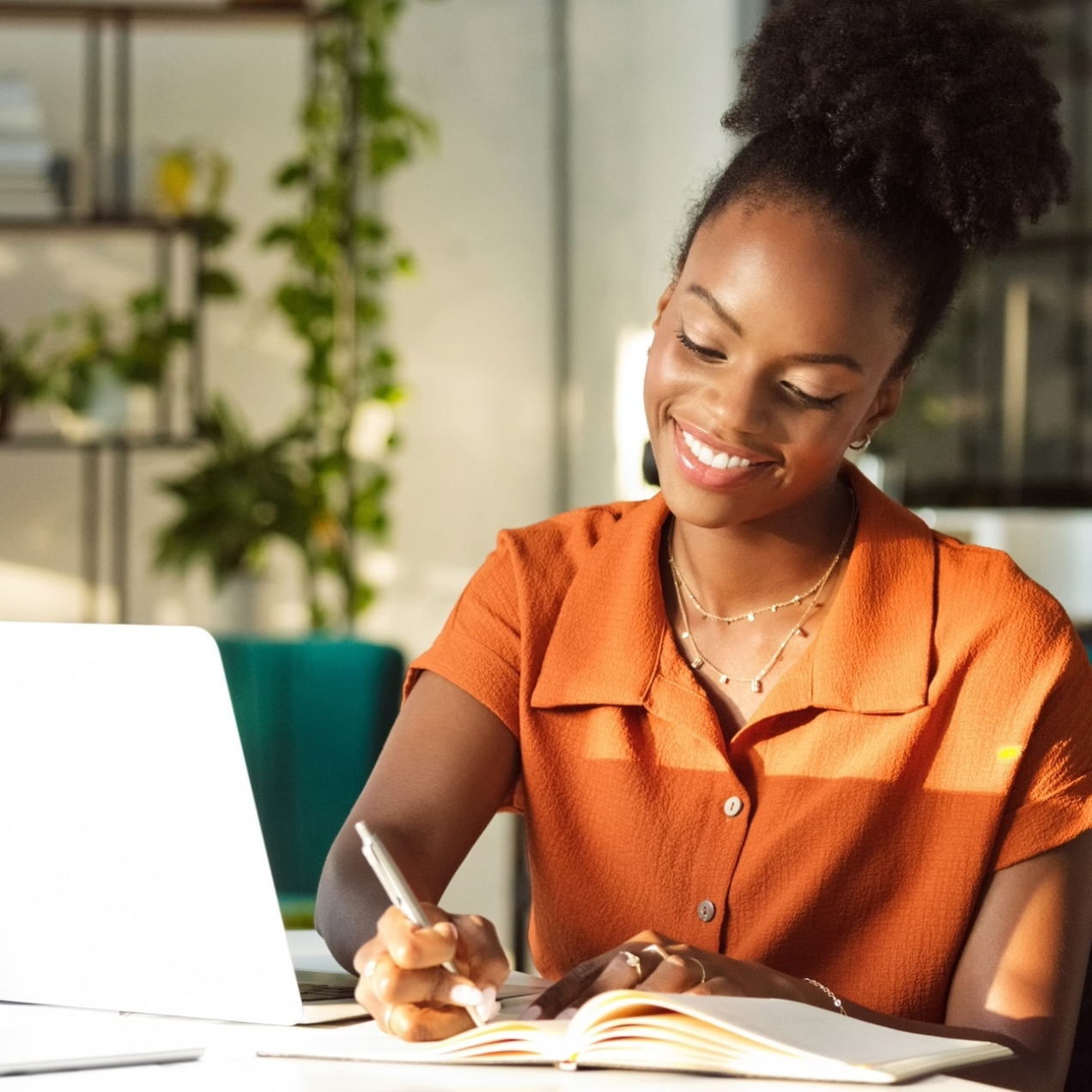 A Black woman wearing an orange blouse smiles while sat at a desk writing in a book with a laptop beside her. She is in a relaxed and trendy office with plants and window.