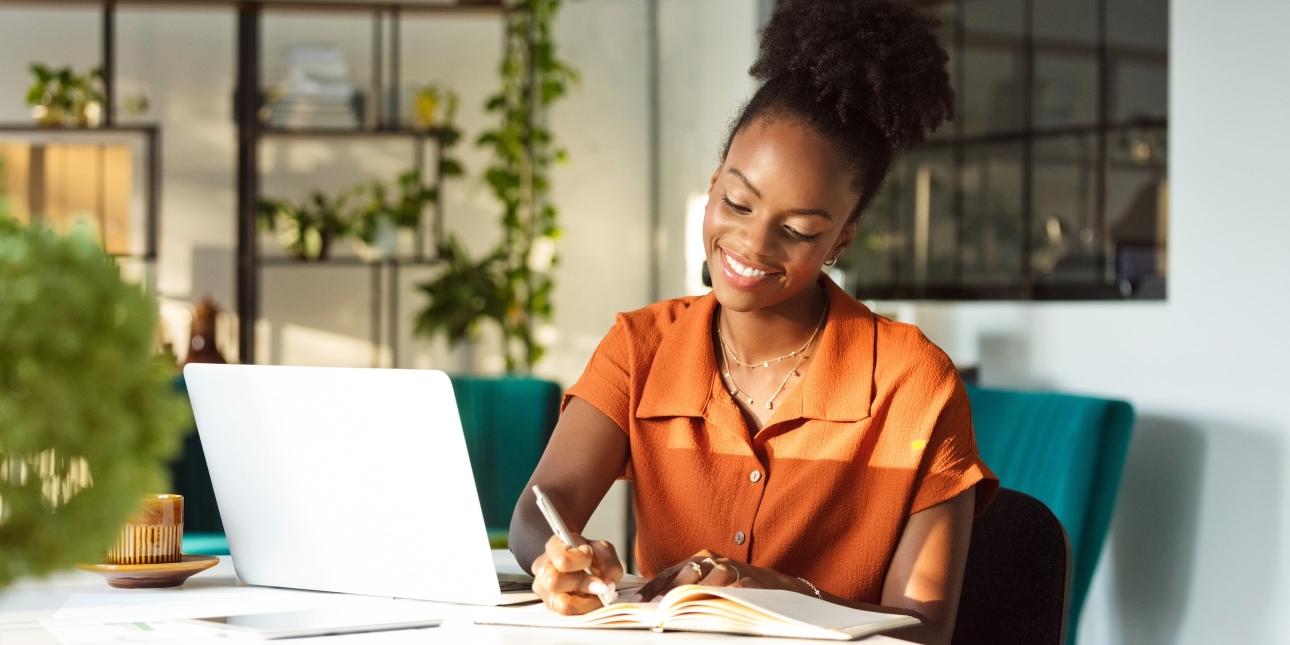 A Black woman wearing an orange blouse smiles while sat at a desk writing in a book with a laptop beside her. She is in a relaxed and trendy office with plants and window.