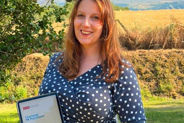 Kirstie Nickson - a white woman with long red hair, who is wearing a white spotted blue dress - holds a framed certificate in a sunny garden.