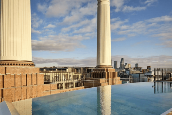 Rooftop infinity pool in front of three of the large chimneys of Battersea power station. The sky is blue with some clouds. The City of London skyline is in the distance