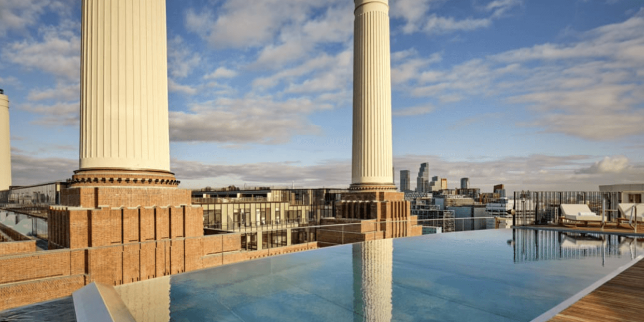Rooftop infinity pool in front of three of the large chimneys of Battersea power station. The sky is blue with some clouds. The City of London skyline is in the distance