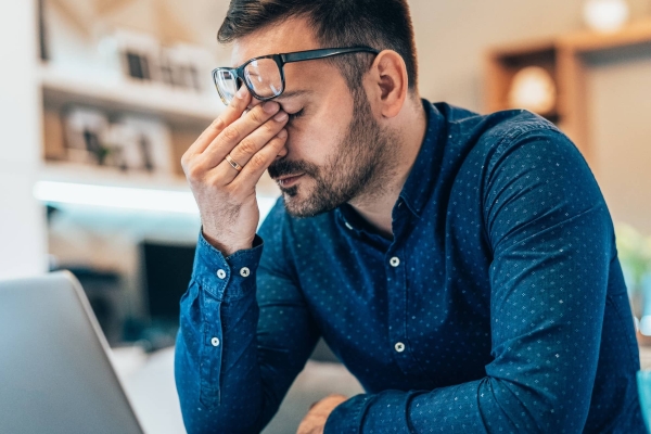 A white man with dark hair and blue shirt sits in front of his laptop. He is rubbing his eyes with his spectacles raised on to his forehead