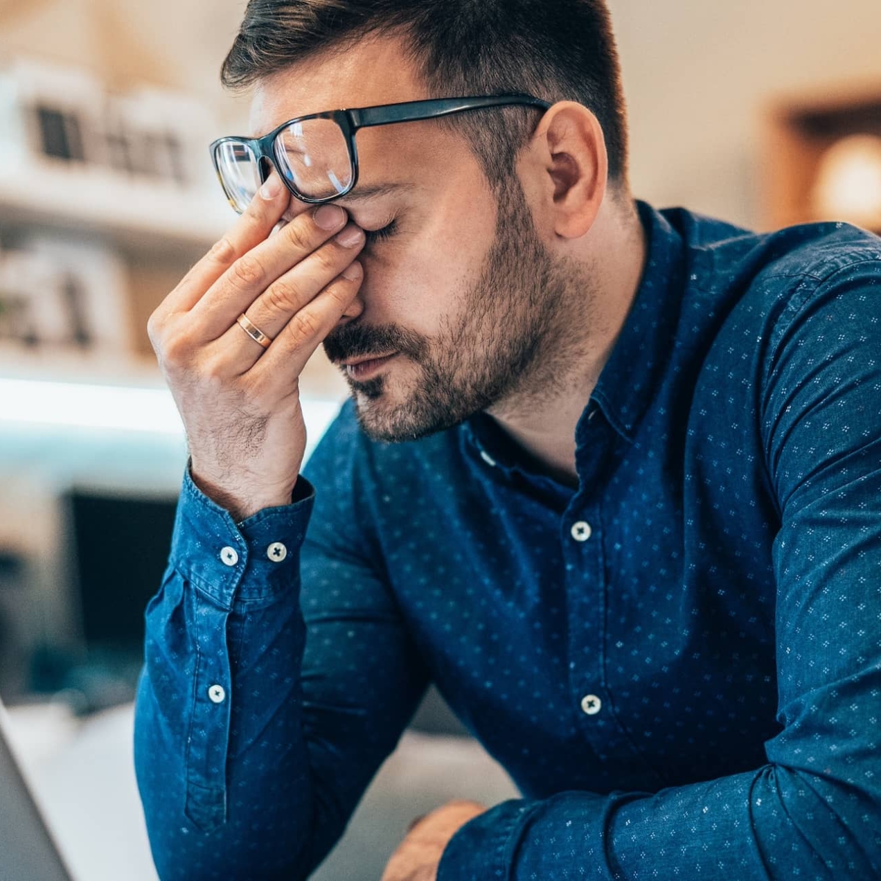 A white man with dark hair and blue shirt sits in front of his laptop. He is rubbing his eyes with his spectacles raised on to his forehead