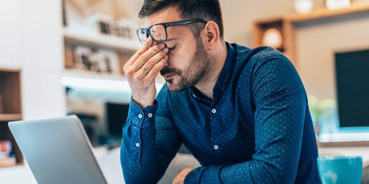 A white man with dark hair and blue shirt sits in front of his laptop. He is rubbing his eyes with his spectacles raised on to his forehead