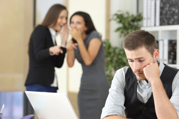 A white man rests his cheek on his hand as he stares dispondantly at his office desk while two women in the background whisper and laugh to each other.