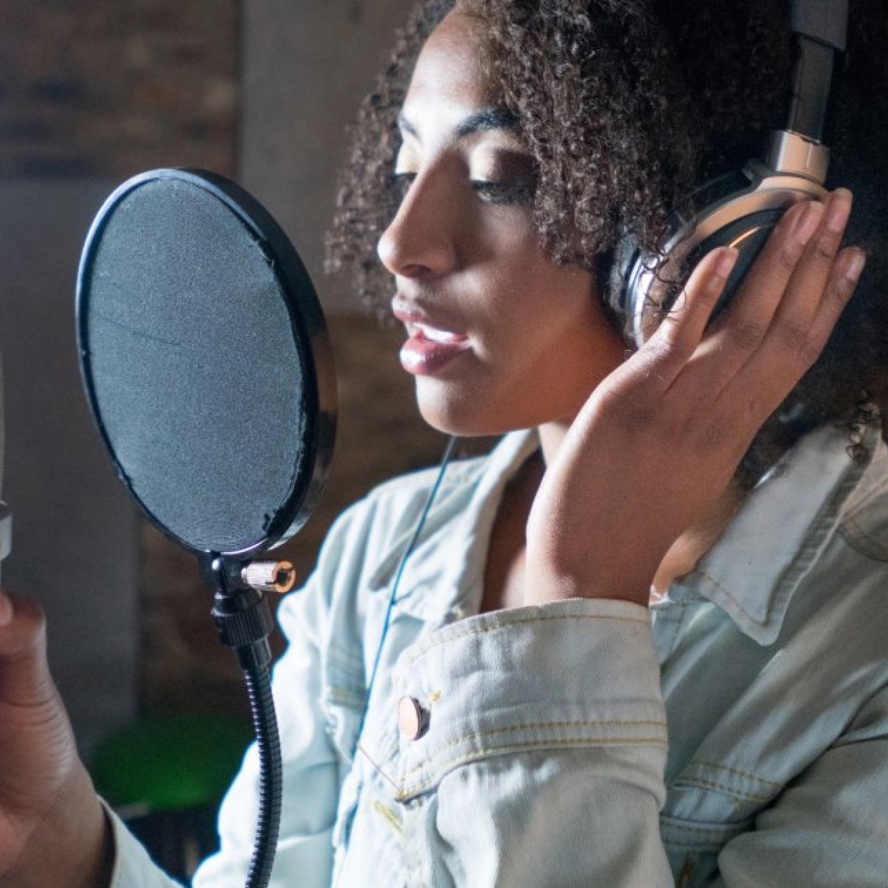 A young Black woman with shoulder length hair, headphones and blue denim jacket sings through a pop filter into a microphone that she is holding
