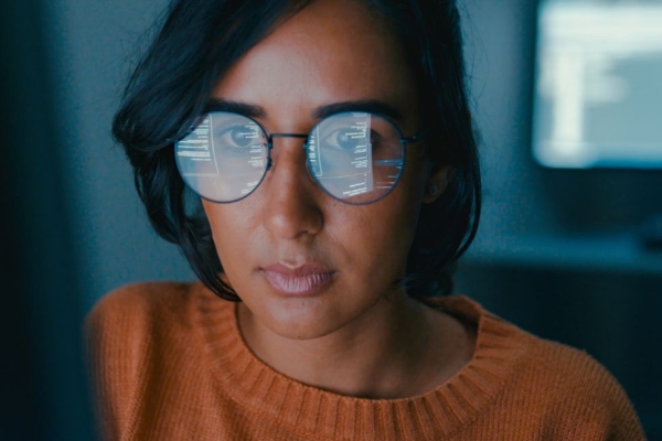 A woman with short dark hair and orange jumper is sat in front of her a computer screen which is reflected in her glasses.