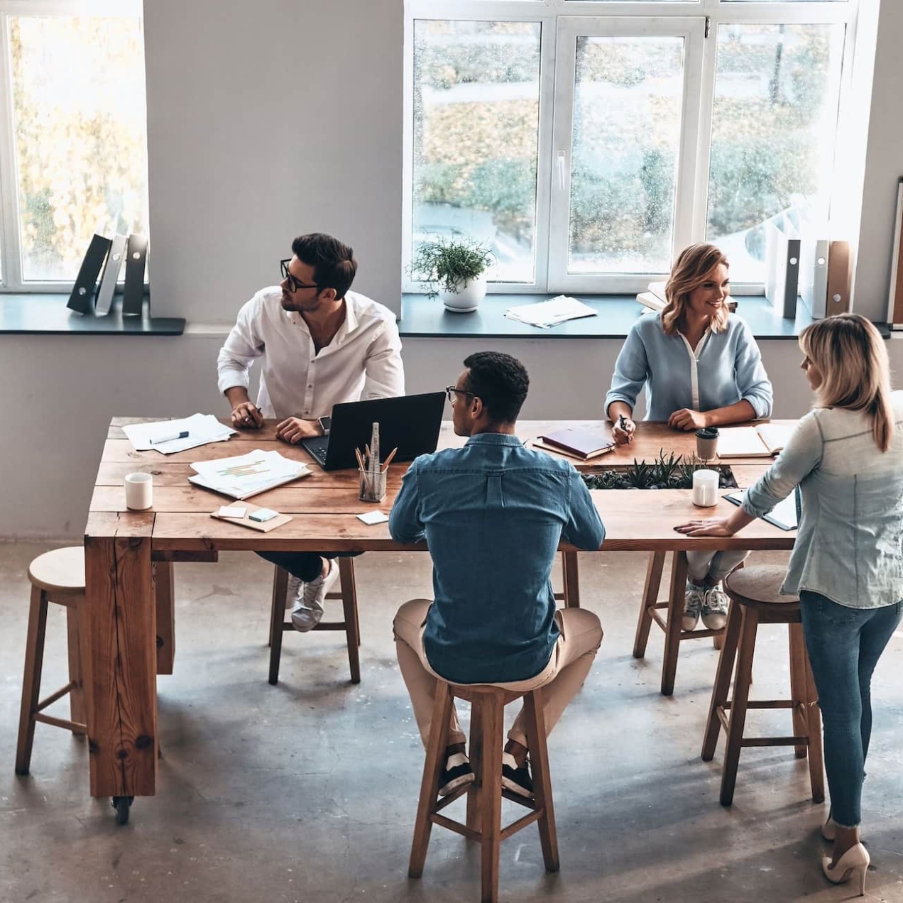 An aerial shot of five men and women of different ethnicities sat in a modern office looking at a white, male colleague with long blond hair as he points to a flip chart of charts