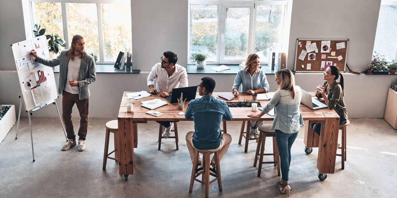An aerial shot of five men and women of different ethnicities sat in a modern office looking at a white, male colleague with long blond hair as he points to a flip chart of charts