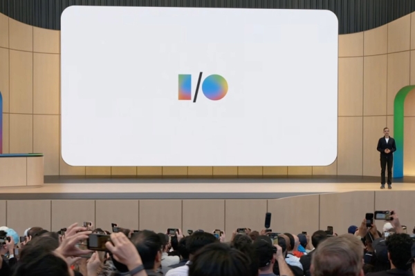 A screenshot from the Google I/O launch event with a shot across the backs of the audience towards a beige stage where a man stands to the right of a screen displaying the I/O logo.