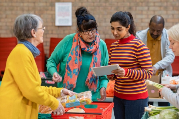Five food bank volunteers of different ages, genders and ethnicities load food into boxes around tables.