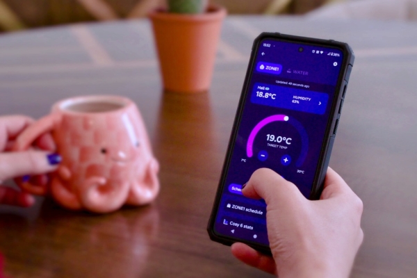 A close up of a white hand holding a mobile phone which displays an energy app. To the left are a pink octopus shaped mug and a small house plant.