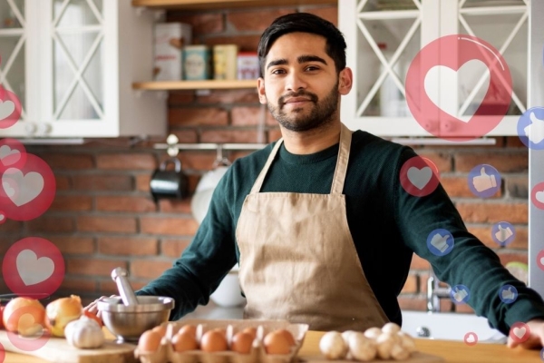 A young Asian man with short hair, who is wearing a beige apron over a green sweatshirt, stands in front of a kitchen counter preparing to cook vegetables. Semi-translucent social media hearts and thumbs up can be seen on the right hand side.