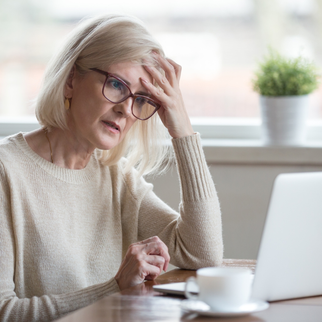 A white woman with grey hair and glasses holds her head with one hand as she stares at the laptop on the desk. She has a look of unease and worry about her.