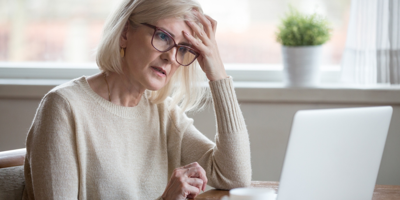 A white woman with grey hair and glasses holds her head with one hand as she stares at the laptop on the desk. She has a look of unease and worry about her.