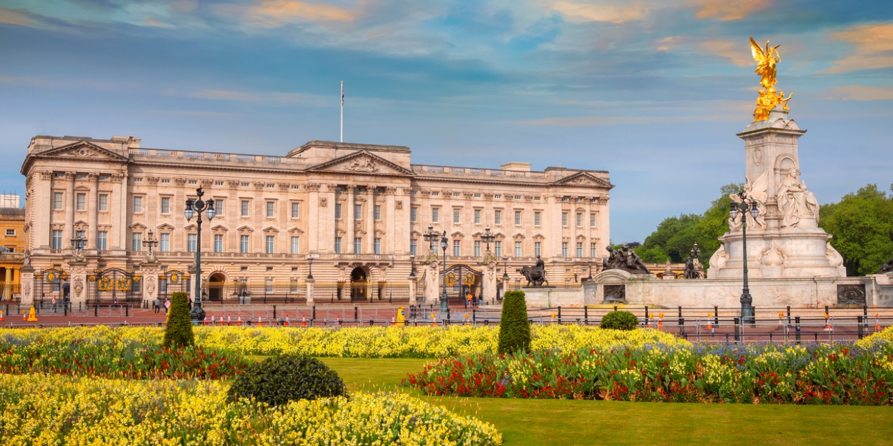 Buckingham Palace against a blue sky. There is green grass and colourful flowers in the foreground.