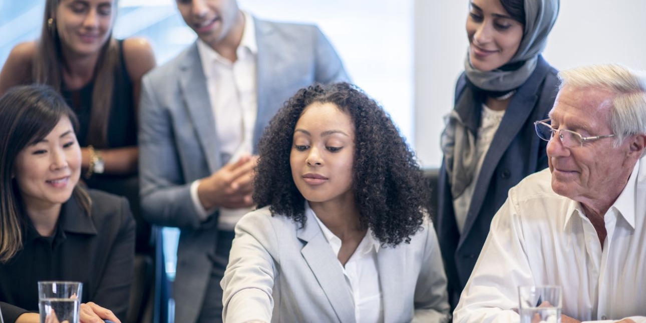 Six smartly dressed and smiling business people gather around a desk looking at paperwork. Four are women, two of which are Asian, one Black and one white. There is one Asian man and one older white man.