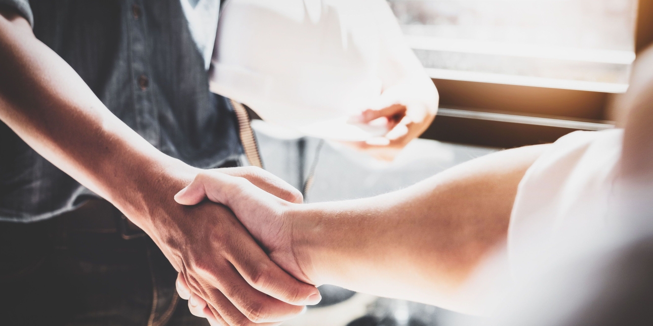 Two unseen figures shake hands, one holding a hard hat and wearing dark clothes