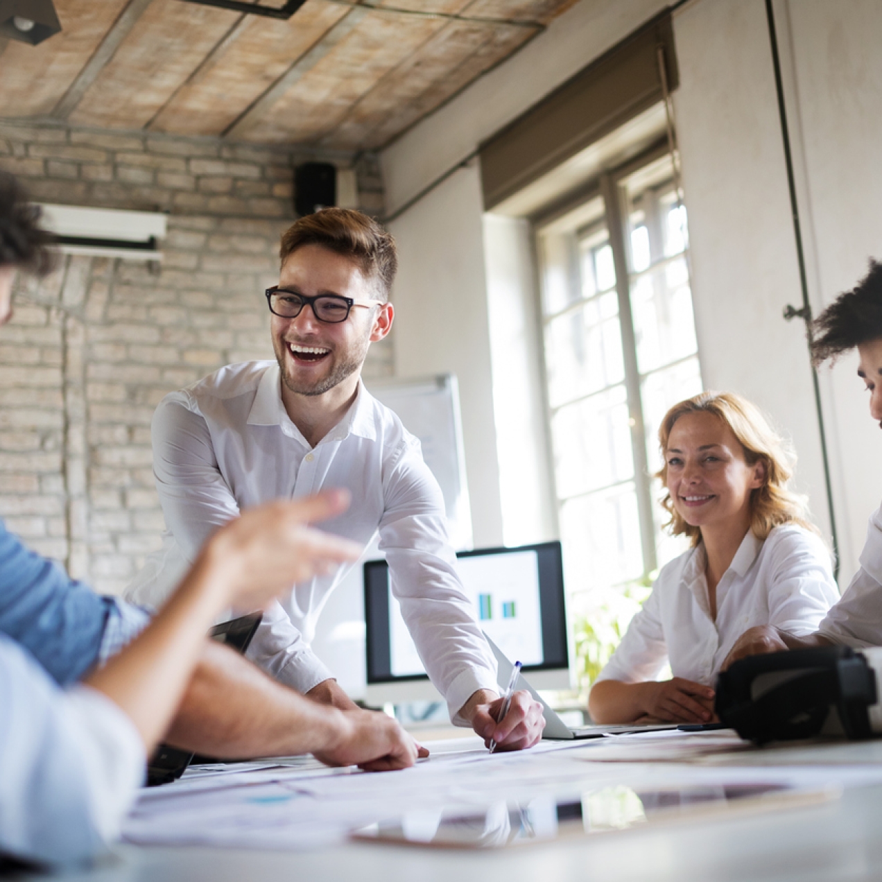 Five business people of different genders and ethnicities smiling around a table in a contemporary office with bare brick walls