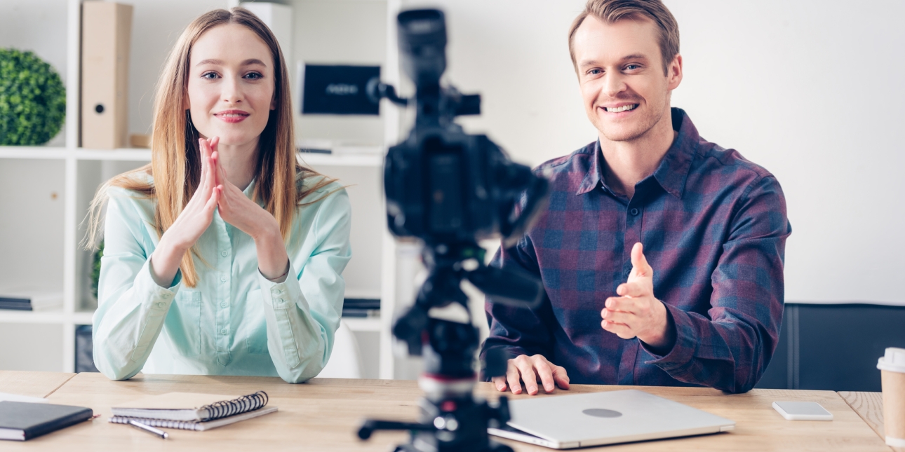 Two business colleagues looking at a video camera. The woman on the left is white with blond hair and smiling. She is holding her palms together with fingers facing upwards. The man on the right is also blond and smiling. He gestures to the camera. T