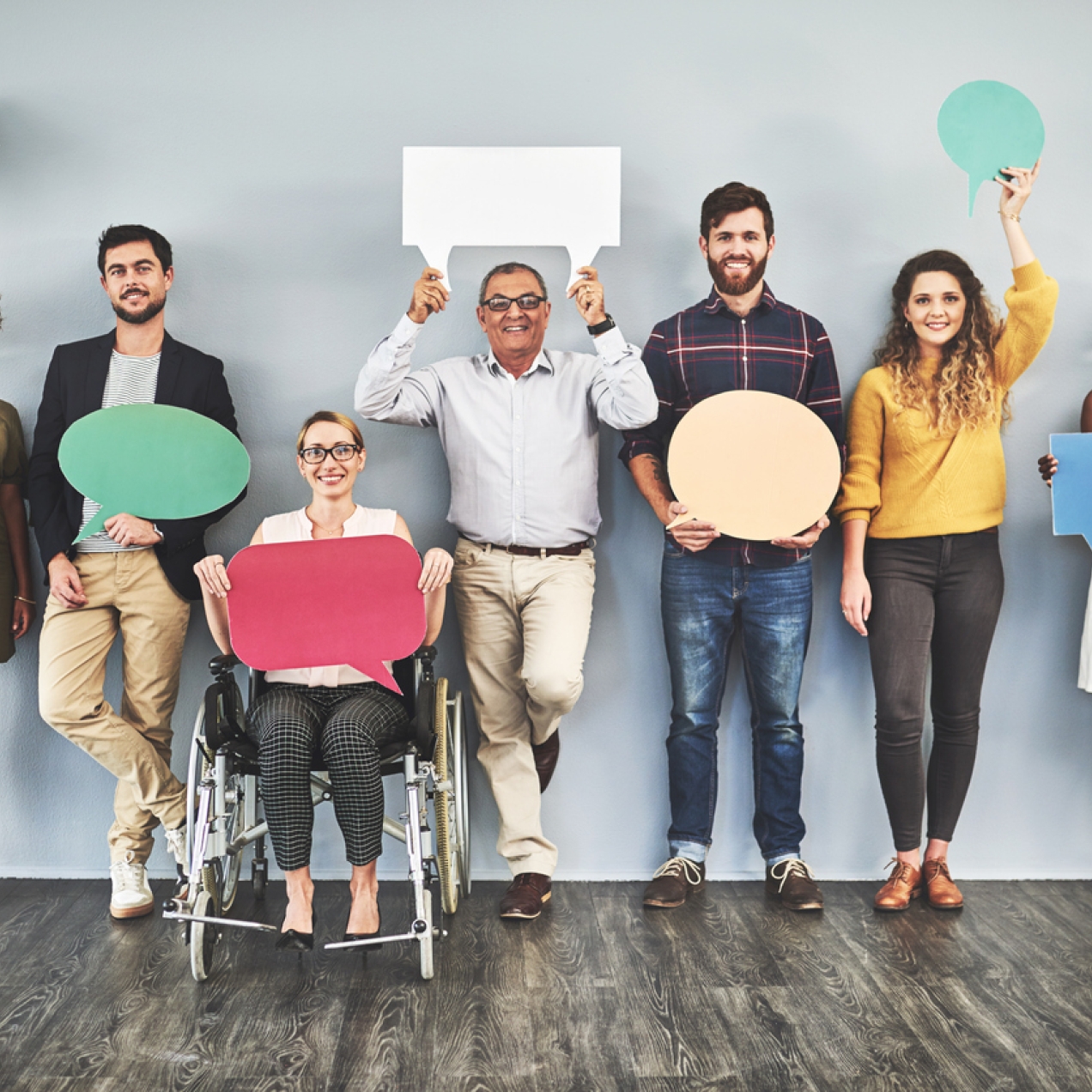 Seven people holding large pieces of card shaped like speech bubbles. Six of the group are standing, one is sat in a wheelchair. The group includes people of different ages, genders and race.