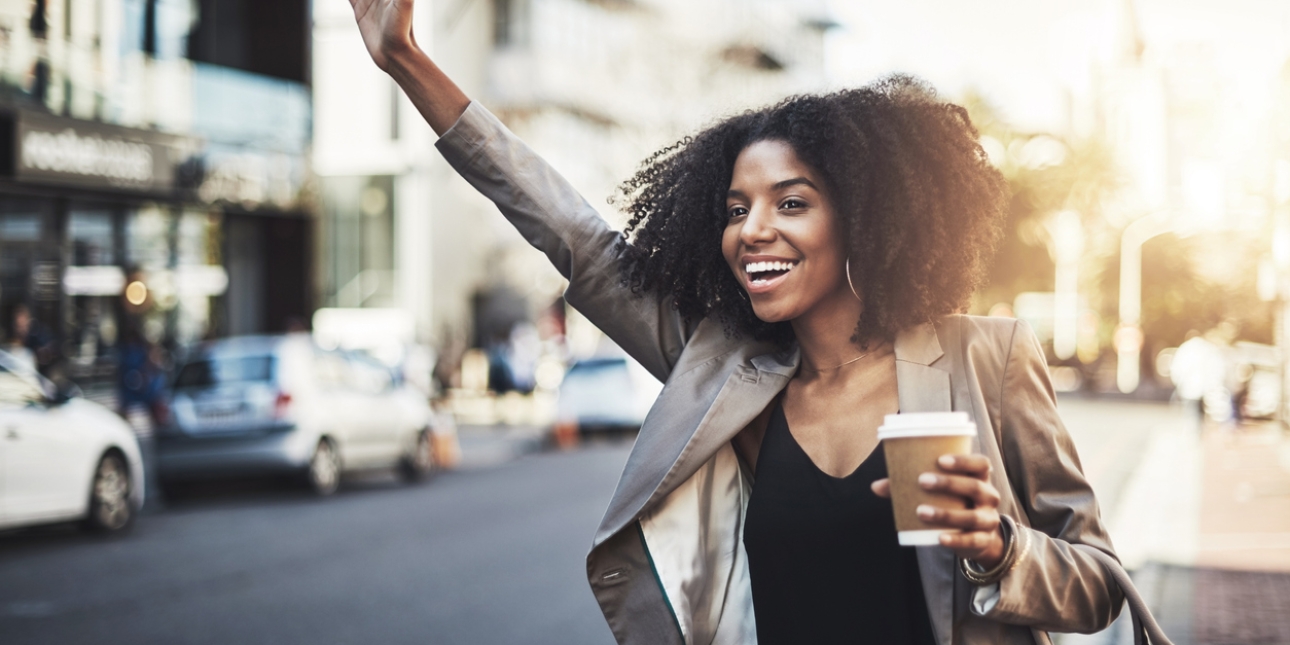 A smiling black woman in a suit jacket is hailing a cab from the side of a city street. Her right hand is raised and the other is holding a coffee cup. She has a matching handbag on this arm.