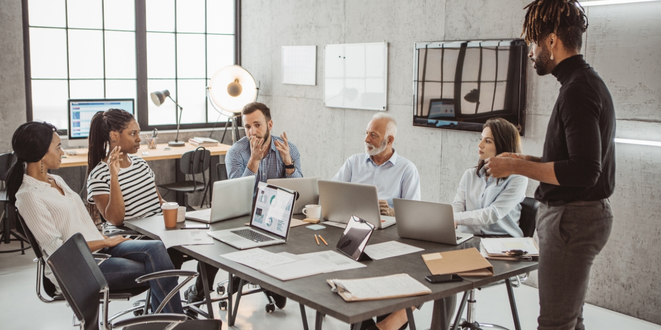 Five business people of different genders, ethnicities and ages sat around a board table. A man is stood.