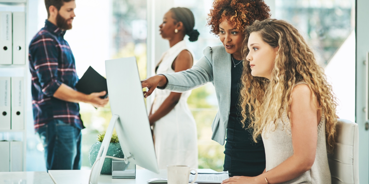 A Black woman in a blazer points at a computer screen of a white woman sat a desk. In the background are a white man and a Black woman stood having a conversation.