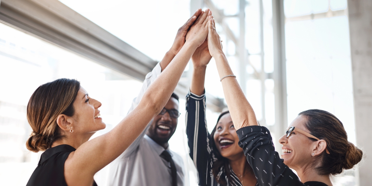 Five business colleagues high-five each other with an up-stretched hand. They comprise of two white women, two black women and one black man. All are dressed in smart-casual clothing.