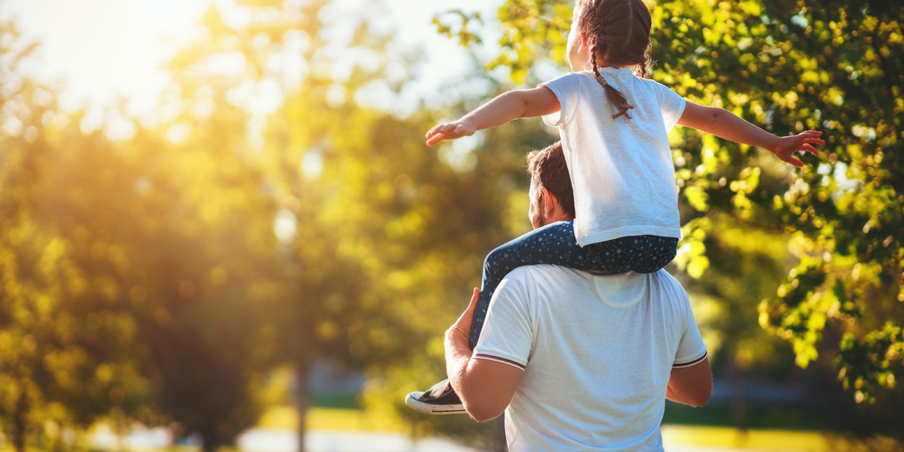 A young girl holds her hands out wide whilst receiving a piggy back from her father during a walk through the park on a sunny day
