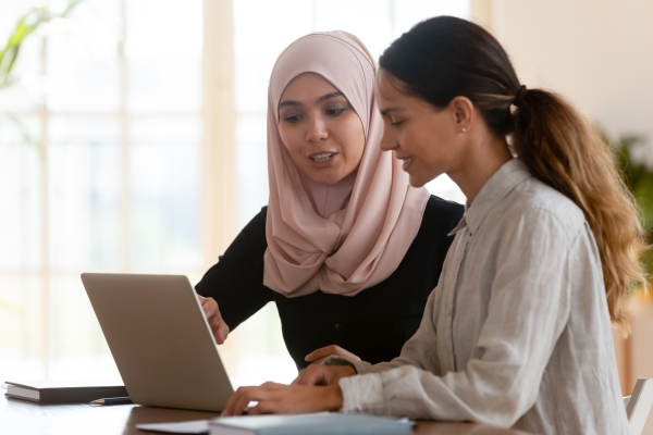 Two women sat around a laptop in a room lit by a large window. The woman on the left, who wears a pink hijab and black long sleeve top, points at the screen. The woman on the right, who has long brown hair tied in a ponytail, wears a long-sleeve grey