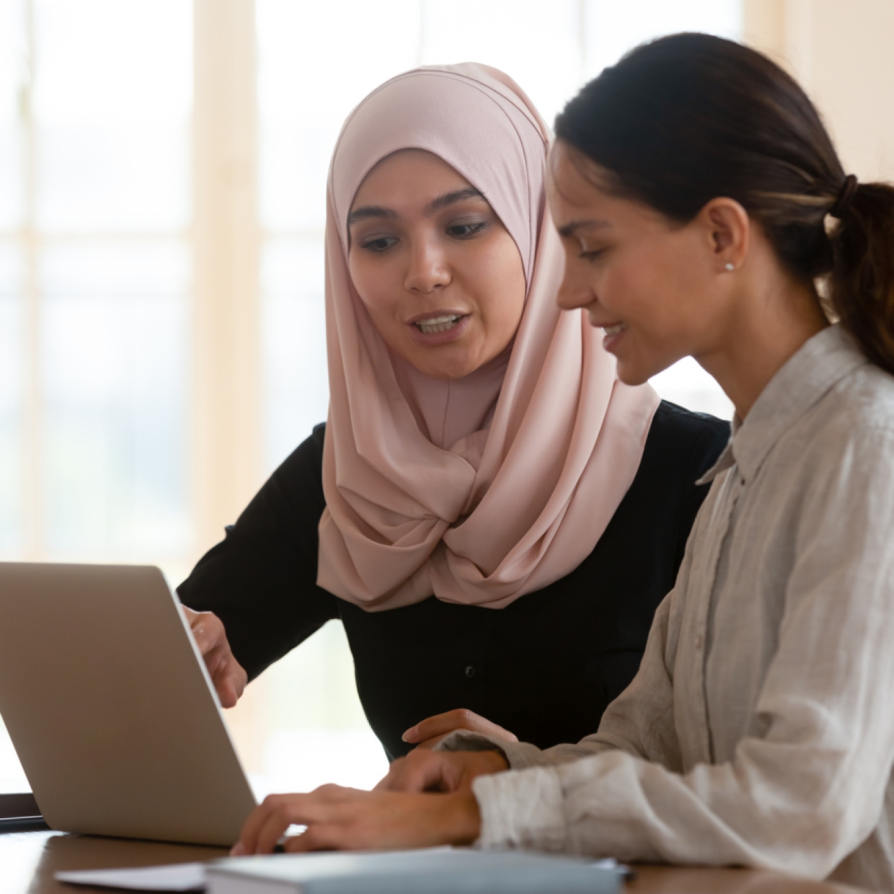 Two women sat around a laptop in a room lit by a large window. The woman on the left, who wears a pink hijab and black long sleeve top, points at the screen. The woman on the right, who has long brown hair tied in a ponytail, wears a long-sleeve grey