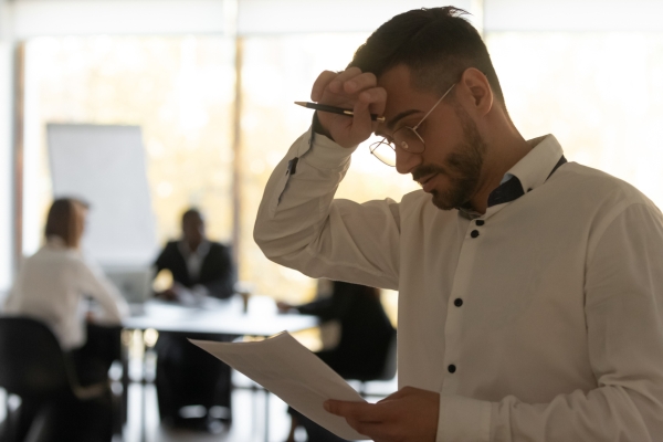 A white man holds his head while reading a piece of paper. Out of the focus in the background are three colleagues sat in a meeting room