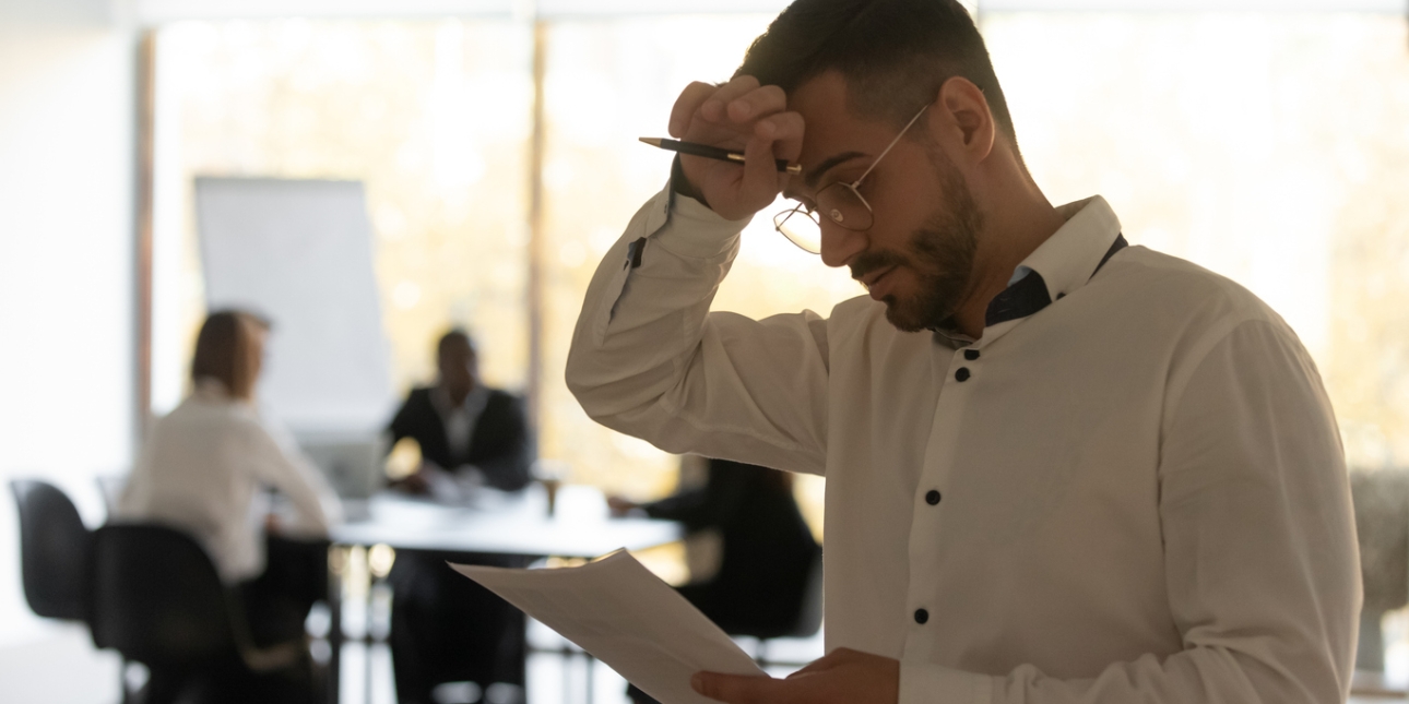A white man holds his head while reading a piece of paper. Out of the focus in the background are three colleagues sat in a meeting room