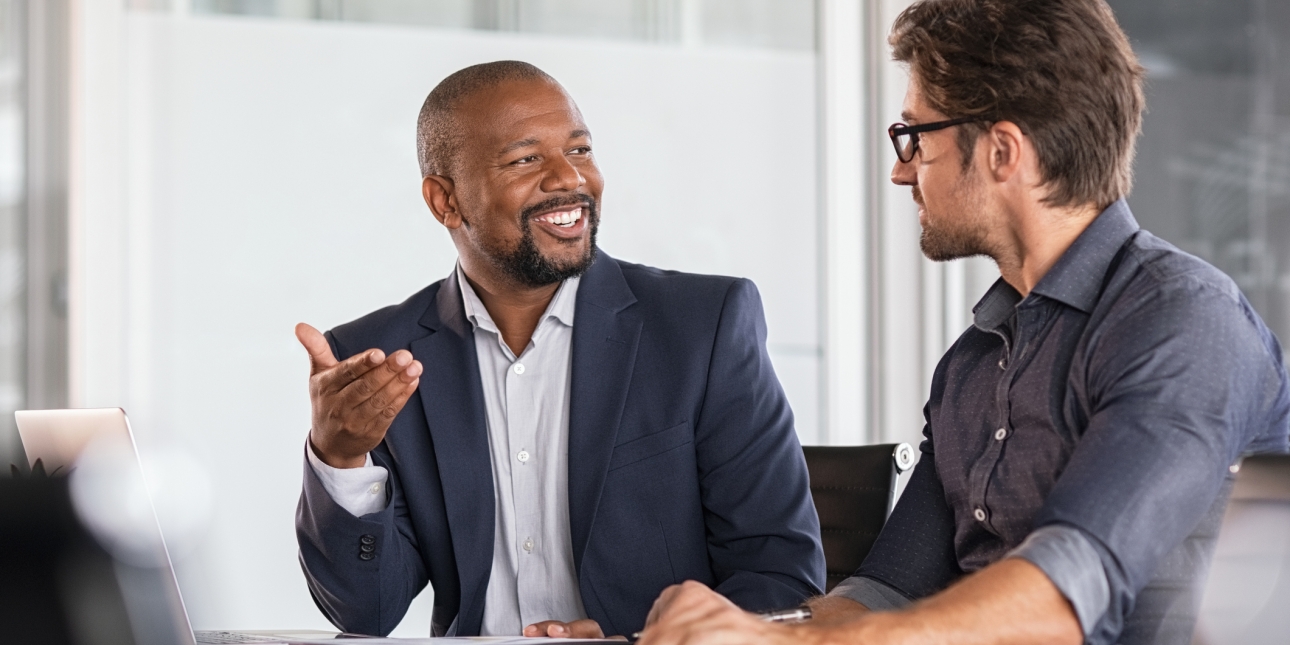 Two colleagues chatting over a desk. On the left is a black male wearing a navy suit jacket and grey scarf who is smiling. On the right, the side profile of a white male colleague wearing a navy shirt and glasses who is listening intently
