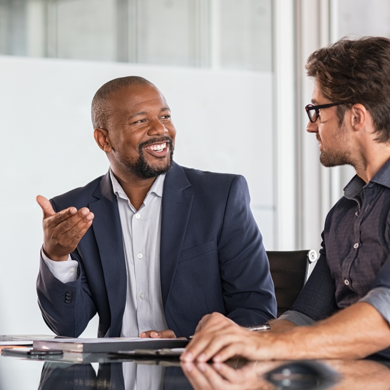 Two colleagues chatting over a desk. On the left is a black male wearing a navy suit jacket and grey scarf who is smiling. On the right, the side profile of a white male colleague wearing a navy shirt and glasses who is listening intently