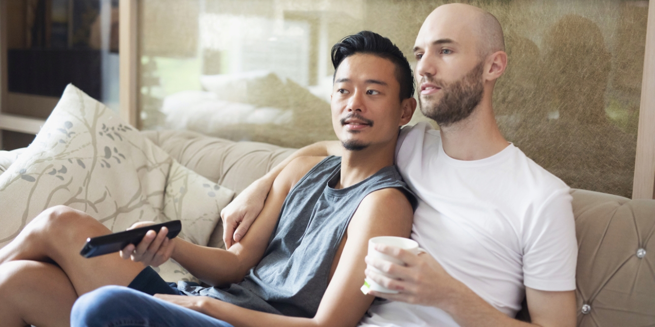 A couple sit cuddling on the sofa while watching television. The man on the left is Asian and wears a vest and short. He is holding a remote control. The man on the right is white with a shaved head and dark beard. He wears a t-shirt and jeans.