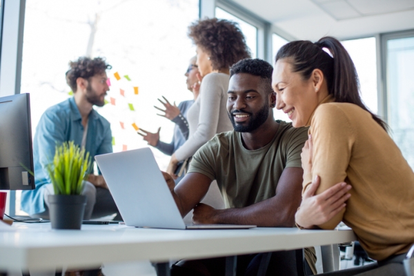 Two colleagues looking at a laptop screen while three colleagues converse in the background