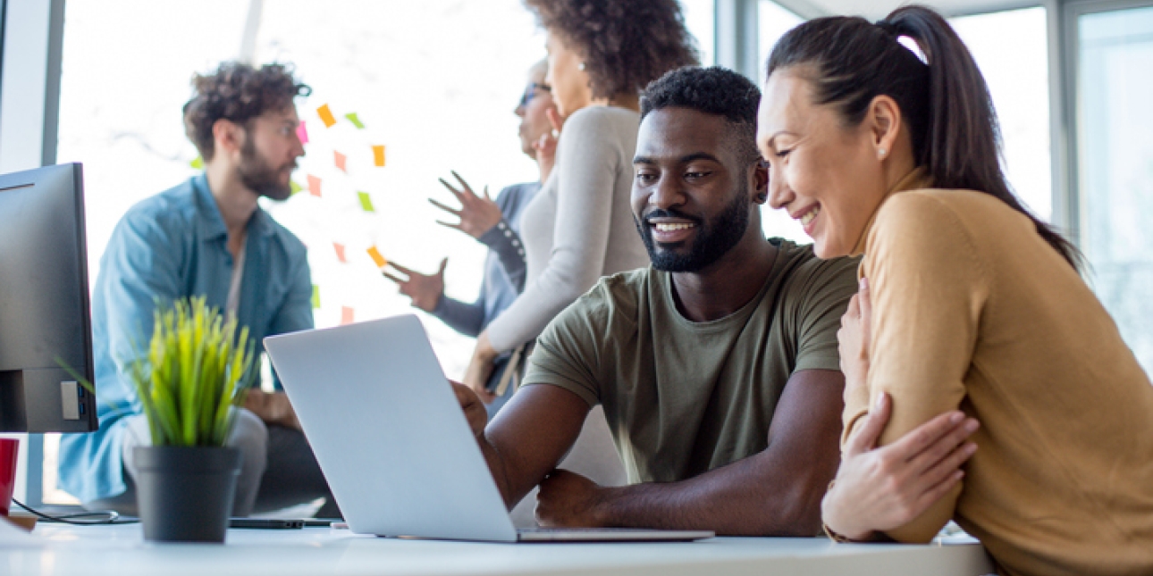 Two colleagues looking at a laptop screen while three colleagues converse in the background