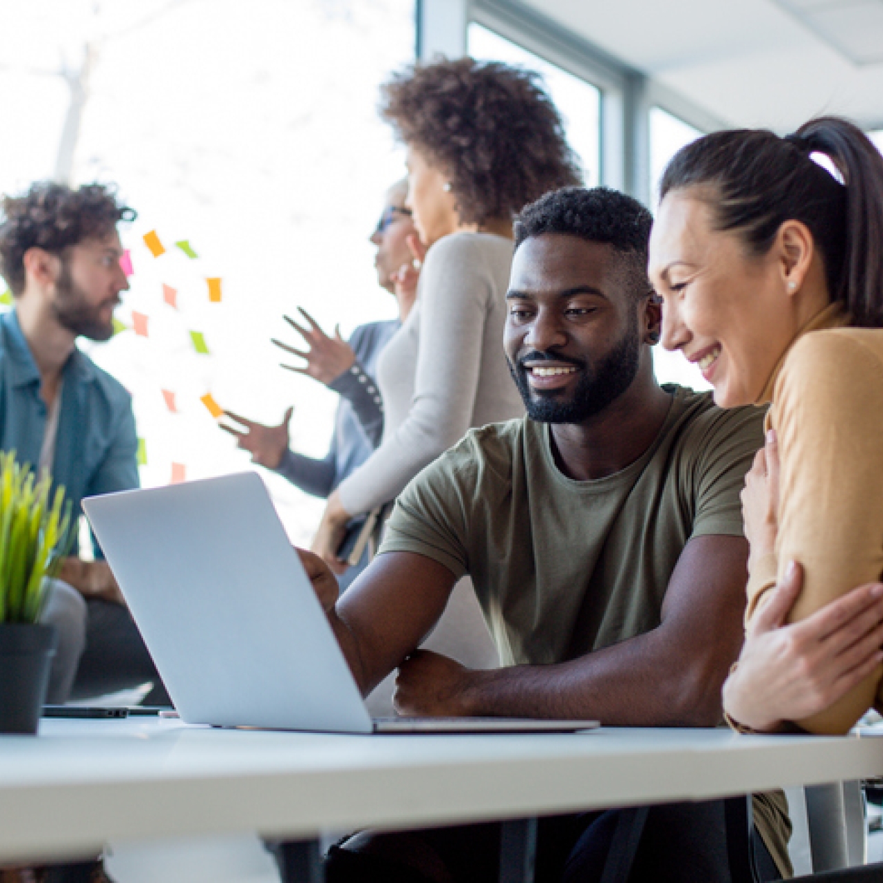 Two colleagues looking at a laptop screen while three colleagues converse in the background